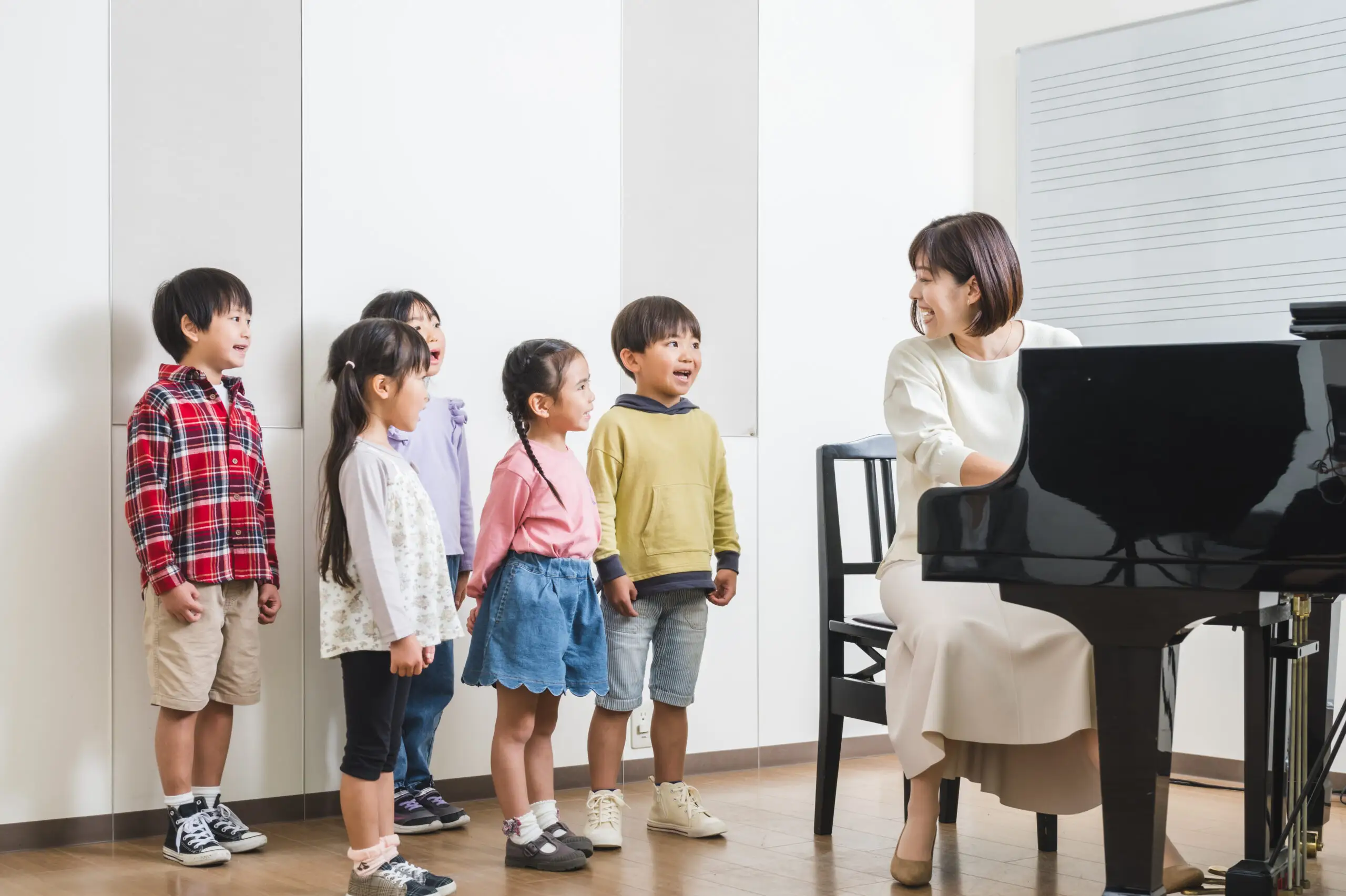 A group of young children standing and singing near a grand piano, while a smiling female music teacher plays the piano and interacts with them in a bright music classroom with a whiteboard in the background.