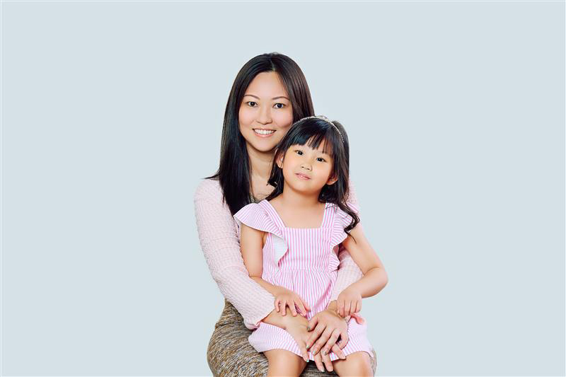 A smiling mother sitting and holding her young daughter on her lap, both looking at the camera. The mother is wearing a light pink sweater and patterned skirt, while the daughter is dressed in a pink and white striped dress with ruffled sleeves, against a plain light blue background.