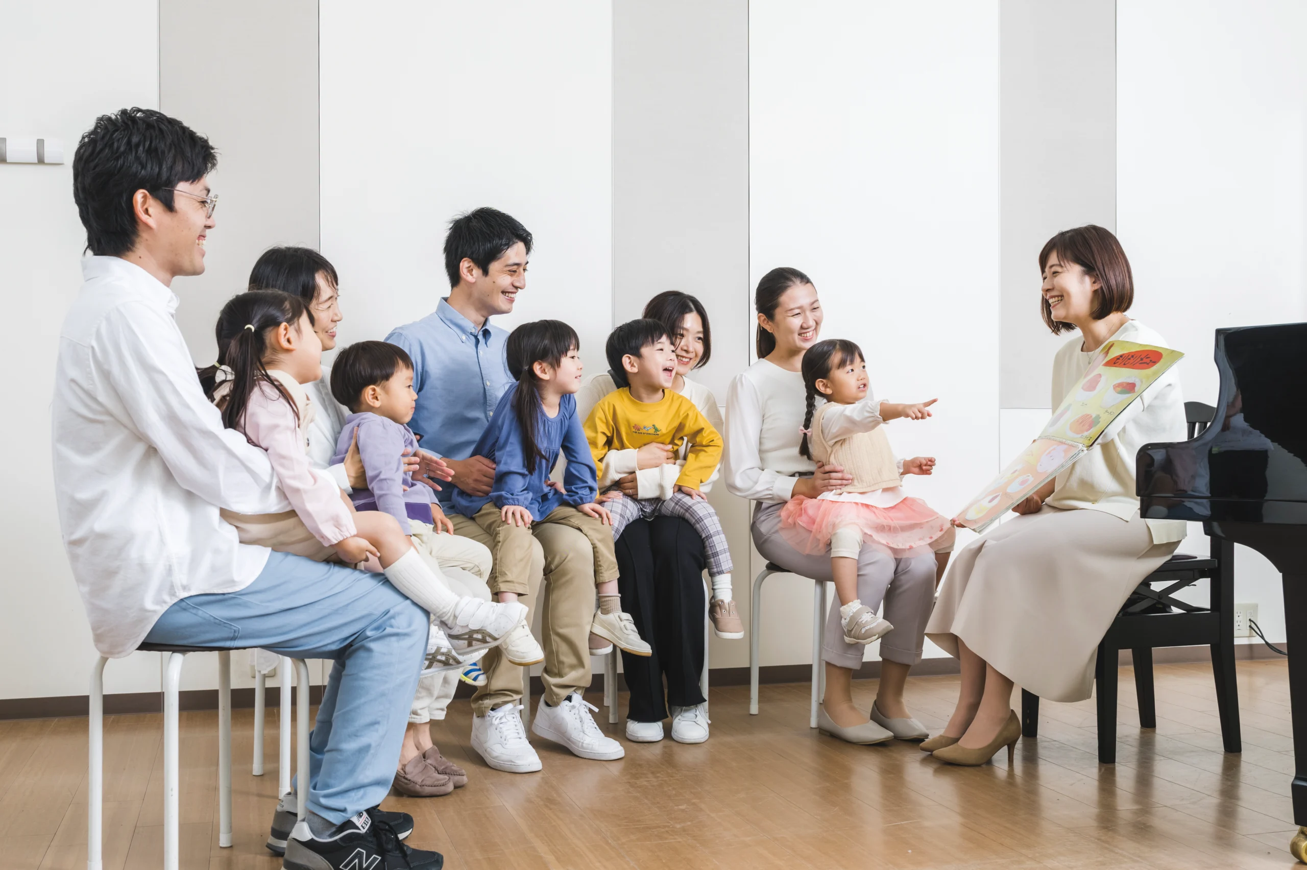 Music teacher engaging a group of young children and parents during an early childhood music class at a Yamaha Music School, with a colorful book and a grand piano in the scene.
